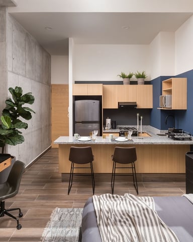 Modern studio apartment with kitchen island, blue accent wall, wood cabinetry, and bed with workspace in foreground