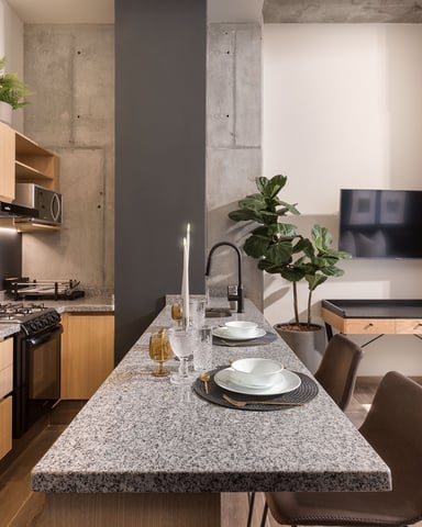 Modern studio apartment with open-plan kitchen, dining counter, living area with TV, and pool table, featuring concrete columns and wood cabinetry.