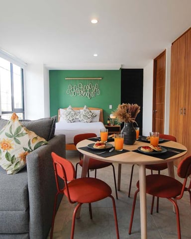 Studio apartment with gray sofa, round dining table with red chairs, and bed with green accent wall in background