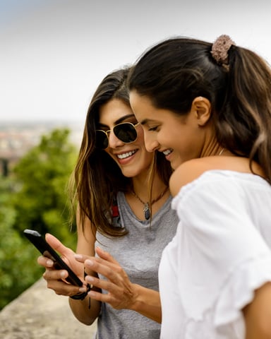 Two women looking at a smartphone on a hilltop overlook with city view and green landscape