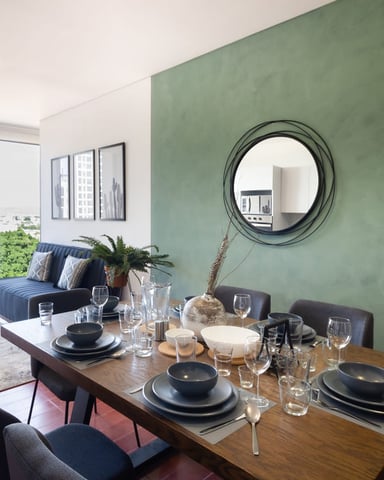 Open-plan dining and living room with sage green accent wall, wooden table set for dining, blue sofa, and city view from balcony