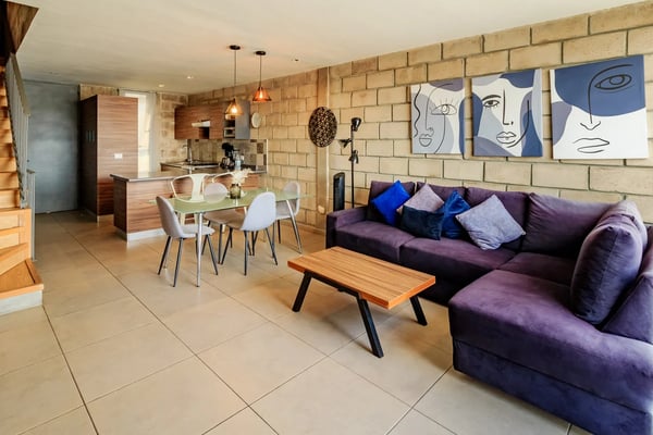 Open-plan living space with purple sectional sofa, dining table, brick wall, and kitchen visible in background