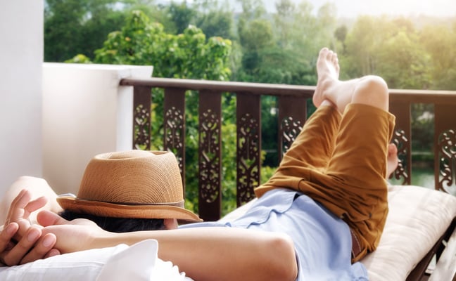 Person relaxing on lounge chair on balcony overlooking forested landscape, wearing straw hat and brown jacket.