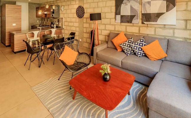 Open-plan living room with gray sofa, red coffee table, orange accents, and brick wall with dining area and kitchen visible in background.