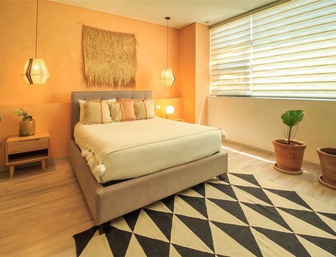 Bright bedroom with orange walls, cream bed with decorative pillows, geometric rug, pendant lights, and potted plants by window with striped blinds.